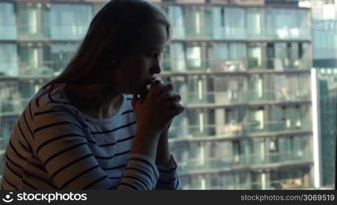 Lonely sad woman with hands close to face sitting by the window with modern cityscape. Loneliness in a big city