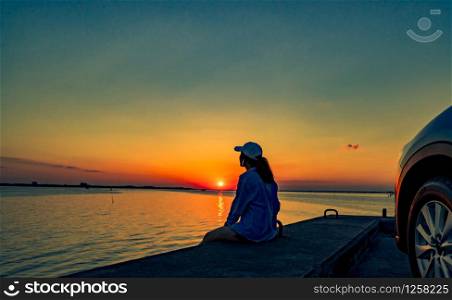 Lonely young woman wear a cap relaxing on the beach alone in front of the car with orange and blue sky at sunset. Summer vacation and travel concept.