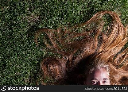 Long blond beautiful hair is spread over summer grass. We can see only half of the face - only pretty eyes. Beautiful young girl is lying on green grass in the evening time.