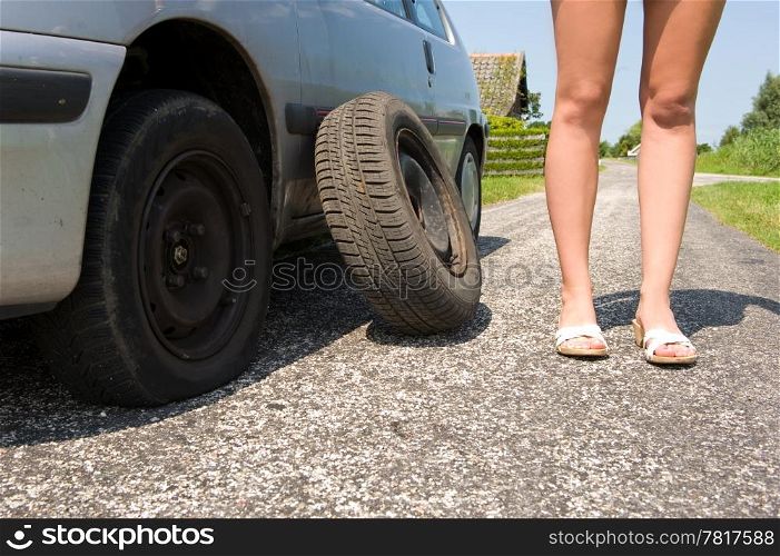 Long legged woman standing next to her car with a flat tire, the spare waiting to be put on