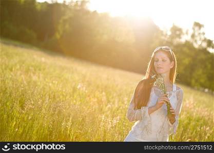 Long red hair woman in romantic sunset meadow holding bunch of flowers