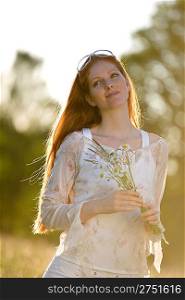 Long red hair woman in romantic sunset meadow holding bunch of flowers