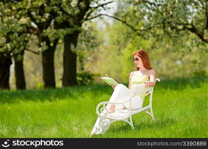 Long red hair woman reading book on white bench in green meadow, shallow DOF