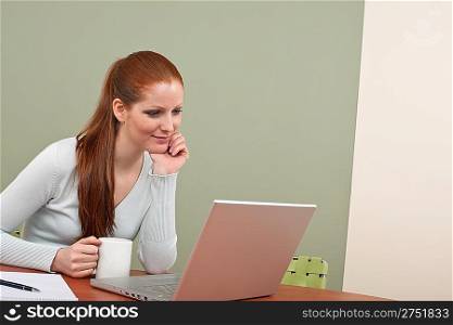 Long red hair woman working at office with laptop sitting at table holding cup of coffee
