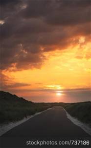 Long street with no cars, through dunes with grass and sand, at sunrise, under an orange cloudy sky, on Sylt island, Germany.