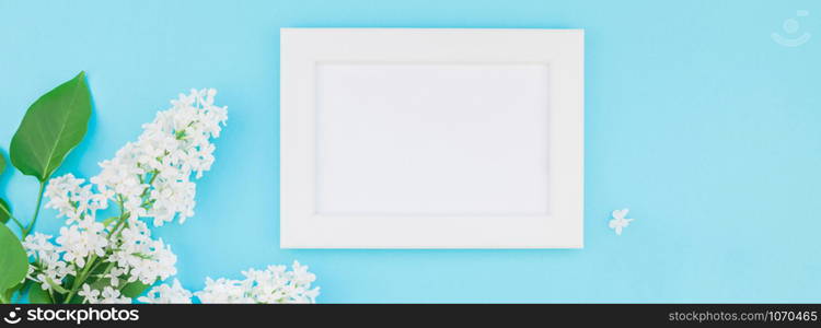 Long wide banner concept top view of blank postcard frame mock up and white lilac flowers petals on pastel blue background with copy space in minimal style, template for lettering, text or design