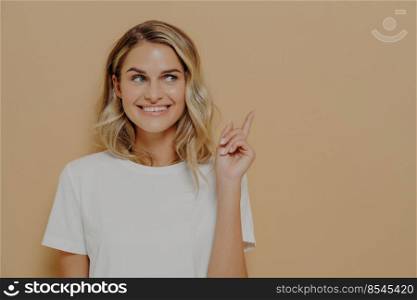 Look at that. Positive young woman customer with blonde hair wearing white tshirt with pleasant smile pointing at copy space on orange background. Advertising and promotion concept. Positive young woman customer with blonde hair wearing white tshirt with pleasant smile pointing at copy space