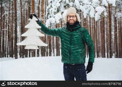 Look at this beautiful fir tree  Cheerful bearded man in fur cap with ear flaps and green anorak, glad to have walk in winter forest, enjoys beautiful trees and nature landscapes. Season concept