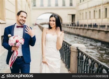 Look at this. Delightful newlyweds show rings on fingers, being glad to have wedding, wear wedding clothes, walk on bridge, have good relationships. People, relations and marriage concept