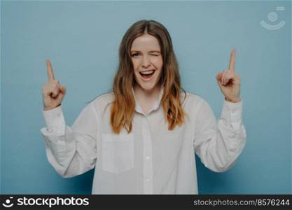 Look here. Happy teenage girl with wide smile in white shirt winking and pointing upwards with her fingers, standing isolated on blue wall. Advertisement concept. Winking happy teenage girl posing against blue wall