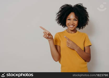 Look here. Studio portrait of beautiful cheerful dark skinned woman dressed in yellow tshirt pointing at left upper corner with forefingers, advertising item while standing against white background. Cheerful dark skinned woman dressed in yellow tshirt pointing at left upper corner with forefingers