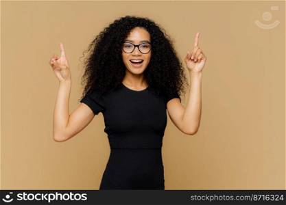 Look upwards. Happy African American woman points above with both index fingers, has slim figure, wears black t shirt, promots object, isolated over brown background. Advertisement and ethnicity