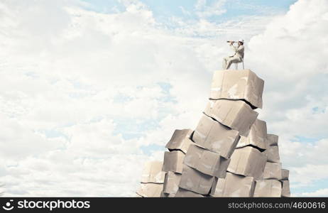 Looking for perspectives. Businessman sitting on big stack of carton boxes and looking in spyglass