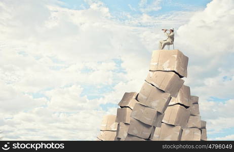 Looking for perspectives. Businessman sitting on big stack of carton boxes and looking in spyglass