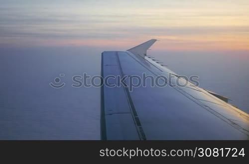 Looking through the illuminator of flying plane at sunset. View to the wing, sky and white clouds beneath