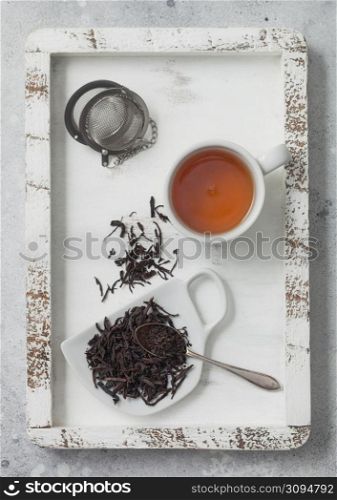 Loose black tea on cup shape ceramic plate with tea ball strainer infuser in wooden box with white ceramic cup. Top view