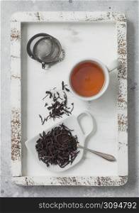 Loose black tea on cup shape ceramic plate with tea ball strainer infuser in wooden box with white ceramic cup. Top view