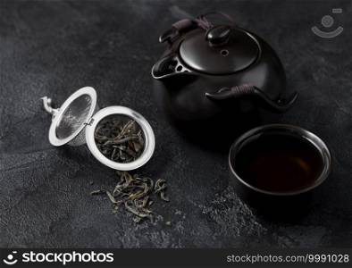 Loose green organic tea in strainer infuser and ceramic teapot and cup on dark background.