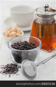 Loose organic black tea with strainer infuser and clear glass teapot with cane sugar and white ceramic cup on light table background.