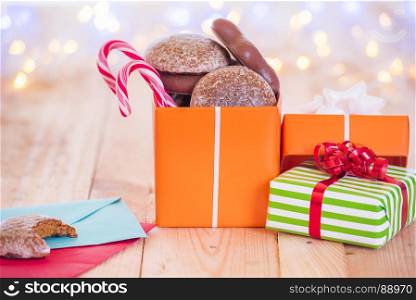 Lots of wrapped presents and an opened gift full with gingerbread and candies, on a wooden table, near envelopes, with bokeh lights in the background.