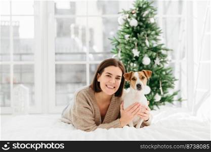 Love and care to pet. Pretty adult woman with short hairstyle, toothy smile, embraces her favourite dog, dressed in warm clothing, lie on comfortable bed against Christmas tree. Winter time.