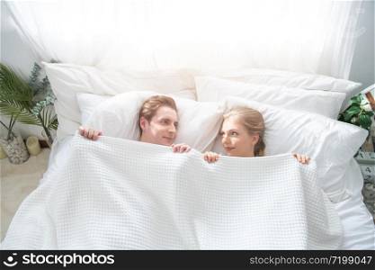 Love and happiness concept .Funny married couple lying in bed and hiding under white blanket, looking at camera with eyes full of joy. Attractive Caucasian man and woman having fun in bedroom.