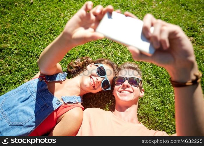 love and people concept - happy teenage couple in sunglasses lying on grass and taking selfie on smartphone at summer. happy couple taking selfie on smartphone at summer