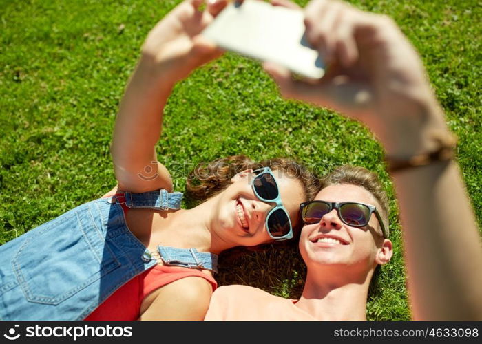 love and people concept - happy teenage couple in sunglasses lying on grass and taking selfie on smartphone at summer. happy couple taking selfie on smartphone at summer