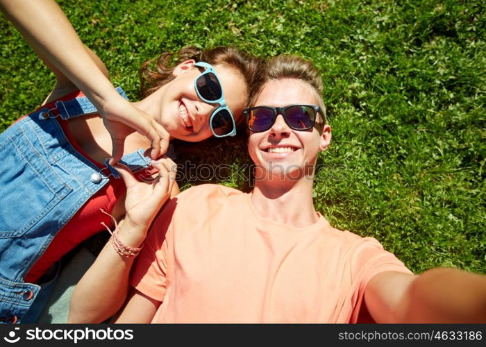 love and people concept - happy teenage couple in sunglasses lying on grass and taking selfie at summer. happy teenage couple taking selfie on summer grass