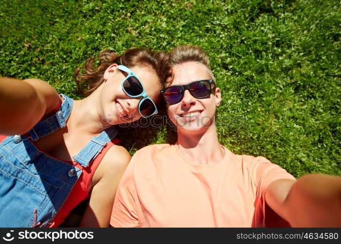 love and people concept - happy teenage couple in sunglasses lying on grass and taking selfie at summer. happy teenage couple taking selfie on summer grass