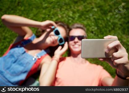 love and people concept - happy teenage couple in sunglasses lying on grass and taking selfie on smartphone at summer. happy couple taking selfie on smartphone at summer