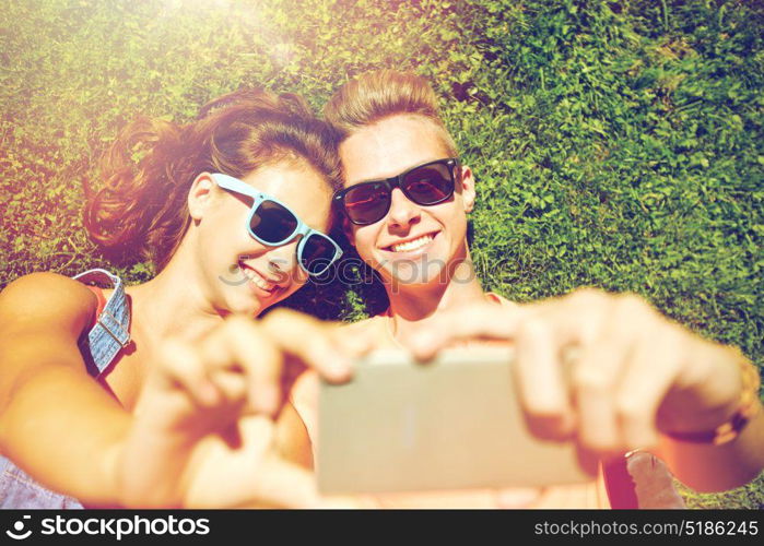 love and people concept - happy teenage couple in sunglasses lying on grass and taking selfie on smartphone at summer. happy couple taking selfie on smartphone at summer