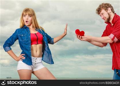 Love and relationship problem. Woman and man young couple in love outdoor, boyfriend giving his girlfriend red heart love symbol, she refusing gift against sky