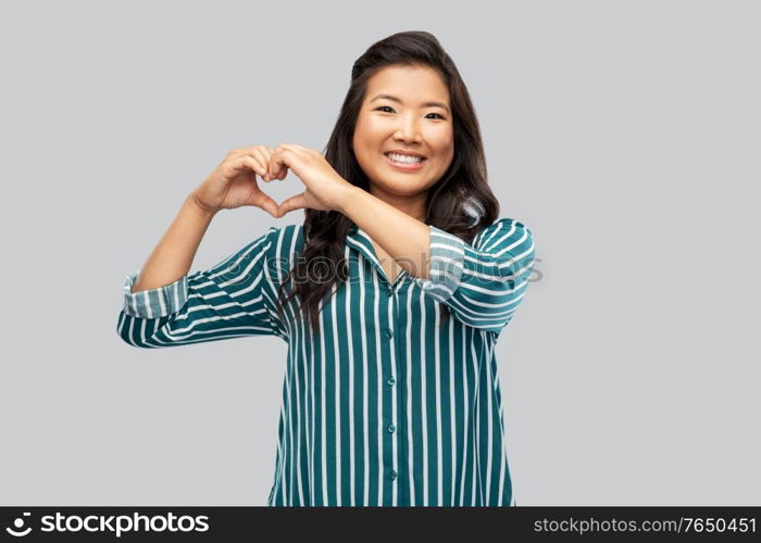 love, charity and valentines day concept - happy asian young woman showing hand heart gesture over grey background. happy asian woman showing hand heart gesture