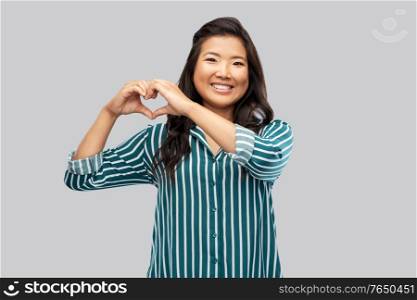 love, charity and valentines day concept - happy asian young woman showing hand heart gesture over grey background. happy asian woman showing hand heart gesture