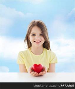 love, charity, childhood and people concept - beautiful little girl sitting at table and holding red heart over cloudy sky background
