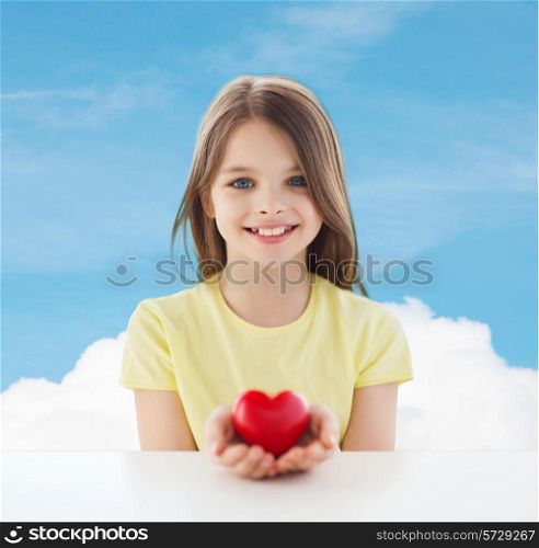 love, charity, childhood and people concept - beautiful little girl sitting at table and holding red heart over blue sky background