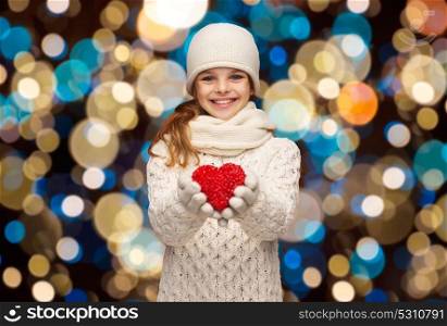 love, charity, christmas, holidays and people concept - happy girl in winter clothes with red heart over lights background. happy girl in winter clothes with red heart