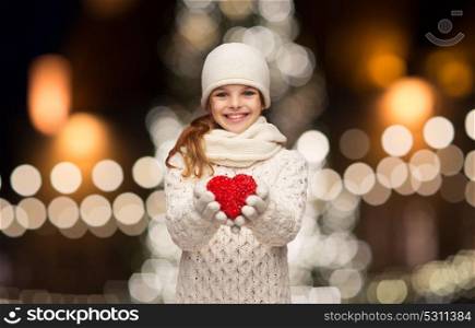 love, charity, holidays and people concept - happy girl in winter clothes with red heart over christmas tree lights background. happy girl in winter clothes with red heart