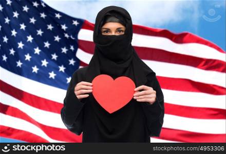 love, charity, immigration, valentines day and people concept - muslim woman in hijab holding red heart over american flag background. muslim woman in hijab holding red heart