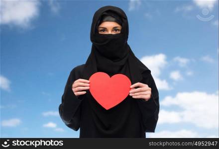 love, charity, valentines day and people concept - muslim woman in hijab holding red heart over blue sky and clouds background. muslim woman in hijab holding red heart
