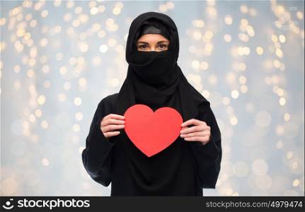love, charity, valentines day and people concept - muslim woman in hijab holding red heart over holidays lights background. muslim woman in hijab holding red heart