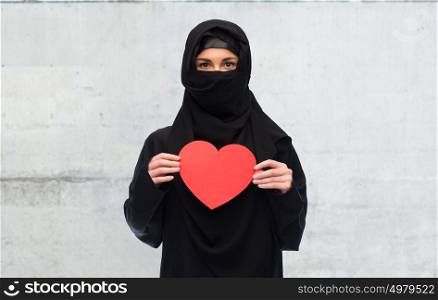 love, charity, valentines day and people concept - muslim woman in hijab holding red heart over gray concrete wall background. muslim woman in hijab holding red heart