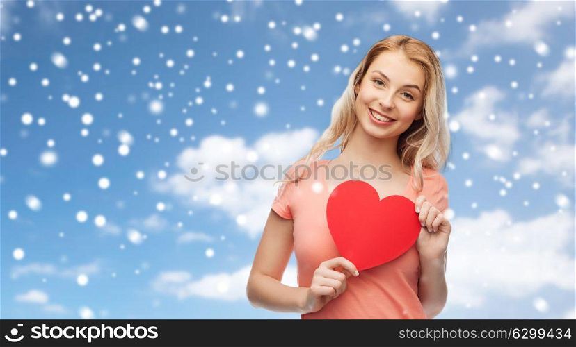 love, charity, valentines day and people concept - smiling young woman or teenage girl holding blank red heart shape over blue sky and clouds background with snow. happy woman or teen girl with red heart shape