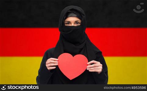 love, charity, valentines day, immigration and people concept - muslim woman in hijab holding red heart over german flag background. muslim woman in hijab holding red heart
