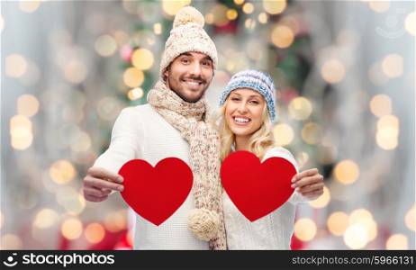 love, christmas, couple, holidays and people concept - smiling man and woman in winter hats and scarf holding red paper heart shapes over lights background