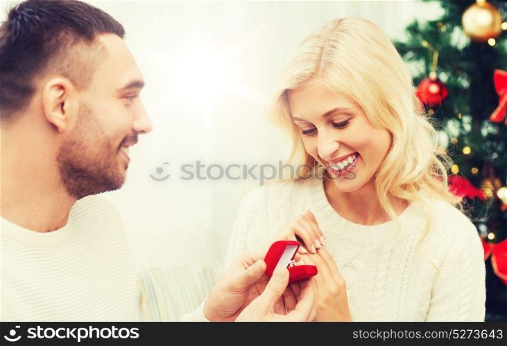 love, christmas, couple, proposal and people concept - happy man giving diamond engagement ring in little red box to woman at home. man giving woman engagement ring for christmas