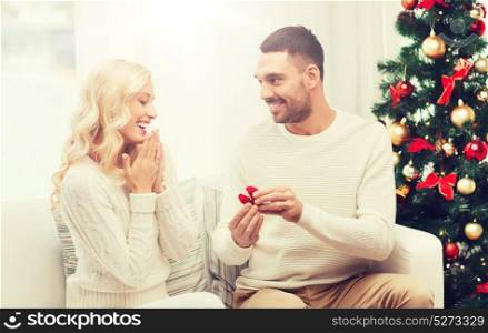 love, christmas, couple, proposal and people concept - happy man giving engagement ring in little red box to woman at home. man giving woman engagement ring for christmas