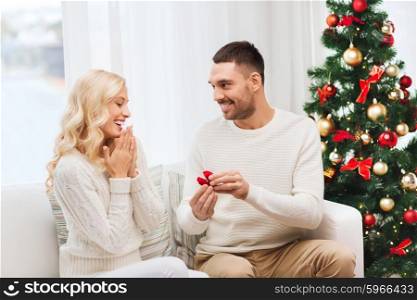 love, christmas, couple, proposal and people concept - happy man giving engagement ring in little red box to woman at home