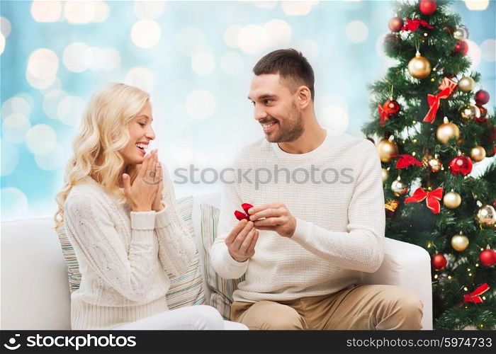 love, christmas, couple, proposal and people concept - happy man giving engagement ring in little red box to woman over blue holidays lights background